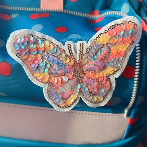 Könnte beinhalten: Ein blauer Rucksack mit einem Punktemuster in Rot und Hellblau, verziert mit einem großen Schmetterlingsaufnäher. Der Schmetterling besteht aus irisierenden, mehrfarbigen Pailletten in Orange-, Rosa- und Goldtönen auf einem weißen Filzhintergrund.