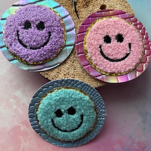 Könnte beinhalten: Drei runde Aufnäher mit Smiley-Gesichtern. Jeder Aufnäher hat ein flauschiges Smiley-Gesicht in Lila, Rosa und Blau, mit schwarzen Augen und einem Lächeln. Sie sind auf einem strukturierten, schillernden Hintergrund mit goldenem Rand angeordnet.