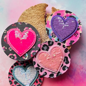Könnte beinhalten: Vier runde Aufnäher mit herzförmigen Pailletten-Designs. Die Aufnäher zeigen rosa, lila und weiße Herzen auf einem Leopardenmuster-Hintergrund. Die Basen sind schwarz oder rosa und haben eine strukturierte Oberfläche.