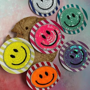 Könnte beinhalten: Sechs kreisförmige Aufnäher mit Pailletten-Smileys in Weiß, Minzgrün, Pink, Gelb, Lila und Orange. Jeder Aufnäher ist auf einer runden Basis mit diagonalen Streifen in passenden Farben montiert, was eine lebendige und strukturierte Sammlung ergibt.