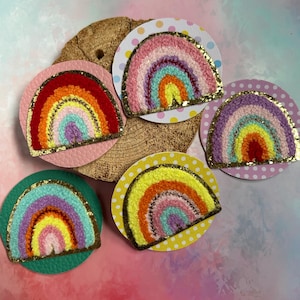 Könnte beinhalten: Fünf kreisförmige Aufnäher mit strukturierten, mehrfarbigen Regenbogenmotiven und goldglitzernden Rändern. Jeder Regenbogen ist auf einer runden Basis mit verschiedenen Mustern und Farben angebracht, darunter Punkte sowie einfarbige Texturen.