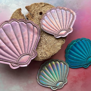 Könnte beinhalten: Vier muschelförmige Aufnäher in Rosa-, Blau- und Regenbogenfarben. Jeder Aufnäher hat ein gesticktes Design und einen gewellten Rand. Sie sind auf einem Stück strukturierten, hellbraunen Materials vor einem rosa-blauen Hintergrund angeordnet.