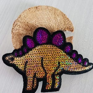 Könnte beinhalten: Ein goldfarbener Pailletten-Dinosaurier-Aufnäher mit schwarzen Rändern und lila Akzenten. Der Aufnäher ist detailliert und hat einen schwarzen Rand. Der Dinosaurier ist vor einem hellen Hintergrund platziert.