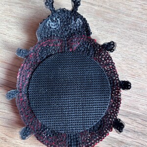 K&ouml;nnte beinhalten: Ein handgefertigter Untersetzer in Form eines Marienk&auml;fers. Der Untersetzer hat eine schwarze Mitte mit einem rot-schwarzen Muster am Rand. Er hat sechs kleine Beine und zwei F&uuml;hler. Er dient zum Schutz von Oberfl&auml;chen vor Hitze und Feuchtigkeit.