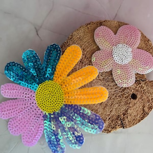 Könnte beinhalten: Zwei paillettenbesetzte Blumendekorationen. Eine große Blume hat rosa, gelbe, blaue und türkisfarbene Blütenblätter mit einem gelben Zentrum. Eine kleinere rosa Blume mit einem weißen Zentrum ist ebenfalls vorhanden. Beide liegen auf einer braunen, strukturierten Oberfläche.