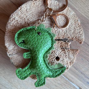 Könnte beinhalten: Ein grüner Dinosaurier-Schlüsselanhänger mit einem goldfarbenen Schlüsselring und Verschluss. Der Dinosaurier besteht aus strukturiertem grünem Material und hat ein genähtes Design. Der Schlüsselanhänger liegt auf einer Holzoberfläche.