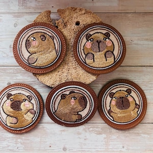 Könnte beinhalten: Set aus fünf runden Untersetzern mit gestickten Capybara-Designs. Jeder Untersetzer hat einen braunen Lederrahmen und einen hellbeigen Hintergrund mit einer Capybara-Illustration. Die Capybaras haben rosa Wangen und sind in verschiedenen Posen.