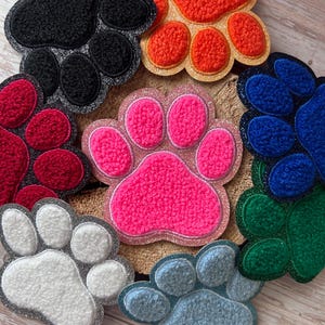 Könnte beinhalten: Eine Sammlung farbenfroher, pfotenförmiger Dekorationsartikel. Die Pfoten sind in verschiedenen Farben erhältlich, darunter Schwarz, Orange, Rot, Pink, Weiß, Blau und Grün. Jede Pfote hat ein strukturiertes, plüschiges Aussehen und ist mit einem glitzernden Rand versehen.