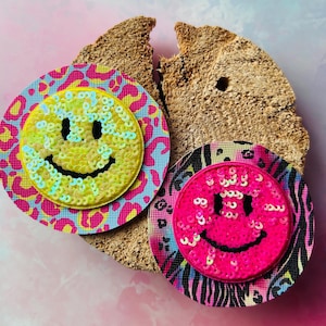 Könnte beinhalten: Zwei paillettenbesetzte Smiley-Aufnäher. Einer ist gelb mit blauen Pailletten, auf einem rosa-blauen Leopardenmuster-Hintergrund. Der andere ist rosa mit rosa Pailletten, auf einem rosa-schwarzen Zebramuster-Hintergrund.
