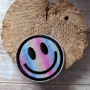 Könnte beinhalten: Ein runder, gestickter Aufnäher mit einem Smiley-Gesicht-Design. Der Aufnäher hat einen schwarzen Rand und eine farbenfrohe, schillernde Mitte mit rosa, blauen und gelben Farbtönen. Der Aufnäher liegt auf einem Holzstück.