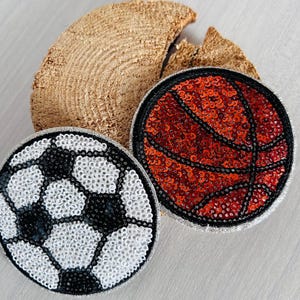 Könnte beinhalten: Zwei paillettenbesetzte Sportball-Aufnäher: ein Fußball mit schwarzen und weißen Pailletten und ein Basketball mit roten Pailletten. Beide haben schwarze Ränder und liegen auf einem Holzstück.