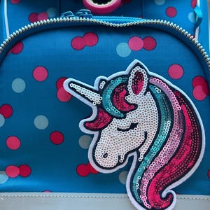 Könnte beinhalten: Ein blauer Rucksack mit weißer Umrandung und Reißverschluss. Der Rucksack hat einen paillettenbesetzten Einhorn-Aufnäher mit weißem Horn, weißem Gesicht und einer bunten Mähne in Rosa-, Türkis- und Blautönen. Der Hintergrund hat ein Tupfenmuster.