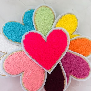 Könnte beinhalten: Eine Sammlung farbenfroher herzförmiger Aufnäher. Die Aufnäher haben eine strukturierte, flauschige Oberfläche in verschiedenen Farben, darunter Blau, Grün, Gelb, Orange, Rosa, Lila und Burgunder. Jedes Herz ist mit goldfarbenen Zierleisten und einem weißen Rand versehen.