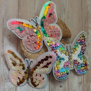 Könnte beinhalten: Drei schmetterlingsförmige Aufnäher, die mit bunten Pailletten verziert sind. Ein Aufnäher zeigt rosa und schillernde Pailletten, ein anderer einen Regenbogen-Farbverlauf und der dritte cremefarbene, beige und braune Pailletten mit schwarzen Fühlern.
