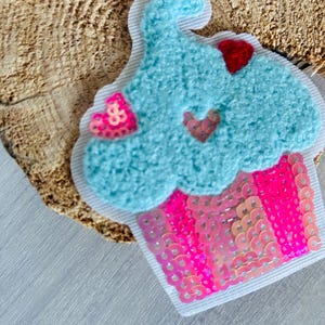 Könnte beinhalten: Ein cupcake-förmiger Aufnäher mit hellblauem, strukturiertem Zuckerguss und einer pink-roten Paillettenbasis. Der Aufnäher hat einen weißen Rand und kleine herzförmige Pailletten.