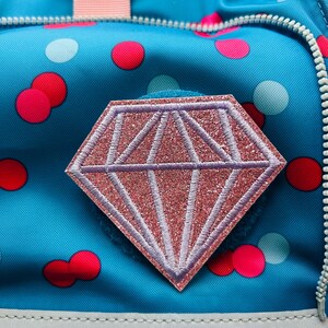 Könnte beinhalten: Ein leuchtend blauer Rucksack mit einem Muster aus roten und hellblauen Punkten. In der Mitte befindet sich ein rautenförmiger Aufnäher mit glitzernder rosa Oberfläche und hellviolettem Stickrand. Ein silberner Reißverschluss und ein rosa Riemen.