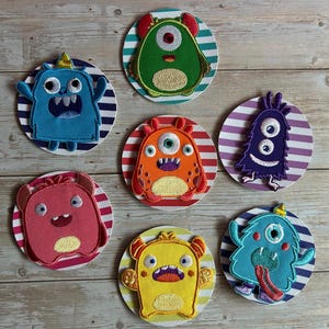 Könnte beinhalten: Sechs kreisförmige, bestickte Aufnäher mit bunten Cartoon-Monstern auf gestreiften Hintergründen. Die Monster sind blau, grün, orange, lila, rosa und gelb, jedes mit einzigartigen Gesichtszügen, Hörnern und Texturen, die mit Garn gestickt sind.