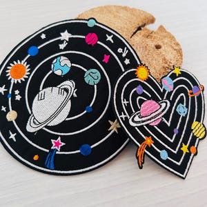 Könnte beinhalten: Zwei gestickte Aufnäher: ein kreisförmiges Design und eine Herzform. Die schwarzen Aufnäher zeigen weiße Umrisse von Planeten, Sternen und Kometen. Die Planeten sind in verschiedenen Farben, darunter Rosa, Blau und Orange. Die Aufnäher sind wahrscheinlich für Kleidung oder Accessoires.