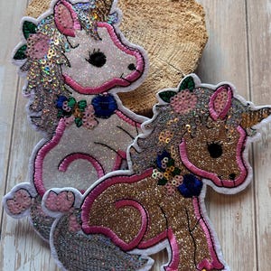Könnte beinhalten: Zwei paillettenbesetzte Einhorn-Aufnäher. Einer ist weiß mit rosa Akzenten, der andere ist goldfarben mit rosa Akzenten. Beide haben florale Details und sind rosa umrandet. Die Aufnäher liegen auf einer Holzoberfläche.