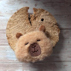 Könnte beinhalten: Ein flauschiges, hellbraunes Bärengesicht mit geschlossenen Augen und einer gestickten braunen Nase und Mund, befestigt an einer rustikalen, kreisförmigen Holzscheibe. Das Bärengesicht ist auf dem Holz zentriert.