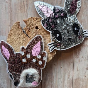 Könnte beinhalten: Zwei aufgenähte Tierkopf-Patches mit Pailletten: ein Reh mit braunen und rosa Pailletten und ein Hase mit grauen und rosa Pailletten. Die Patches befinden sich auf einem Stück Holz mit einer hellbraunen, strukturierten Oberfläche. Der Hase hat schwarze Augen und Schnurrhaare.