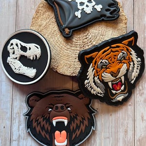 Könnte beinhalten: Eine Sammlung von Aufnähern in Schwarz, Weiß, Braun und Orange. Designs umfassen eine Piratenflagge, einen Dinosaurierschädel, einen brüllenden Bären und einen knurrenden Tiger. Diese Aufnäher sind für Kleidung oder Accessoires geeignet.