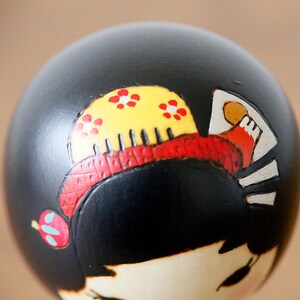 May include: Close-up of a Japanese Kokeshi doll head. The doll has black hair, a yellow comb with red floral accents, and a red headband. The doll's face is pale with black eyes and pink cheeks.