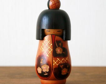 May include: A wooden Japanese Kokeshi doll. It has a black head and a brown body decorated with gold and brown floral and geometric patterns. The doll stands on a wooden surface. The doll is approximately 15 cm tall.