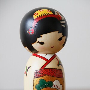 May include: A Japanese Kokeshi doll with a black head and a cream-colored body. The doll's head is adorned with a yellow and red headdress. The doll's kimono is decorated with red, gold, and green patterns. The doll is a traditional Japanese craft.