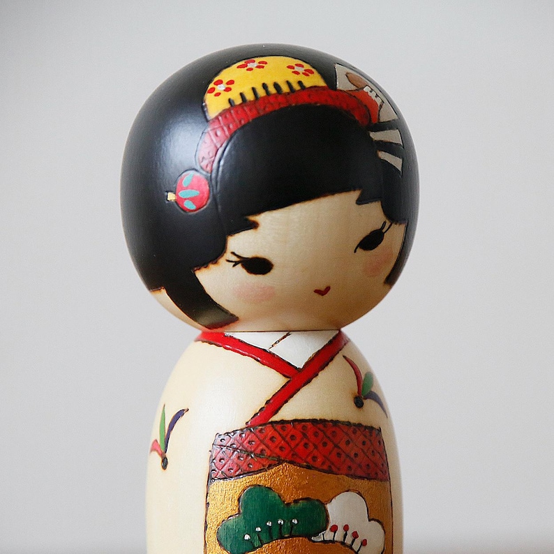May include: A Japanese Kokeshi doll with a black head and a cream-colored body. The doll's head is adorned with a yellow and red headdress. The doll's kimono is decorated with red, gold, and green patterns. The doll is a traditional Japanese craft.