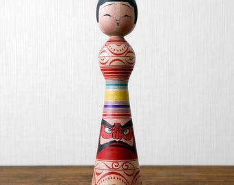 May include: A traditional Japanese Kokeshi doll made of wood. The doll has a smiling face and black hair. It features a red, white, and black design with a geometric pattern. The base of the doll has a fierce face design.