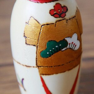 May include: A close-up of a Japanese Kokeshi doll. The doll is cream-colored with gold, red, and green painted details. The design includes a red flower, a green tree, and a white cloud with red accents. The doll has a red band around the top.