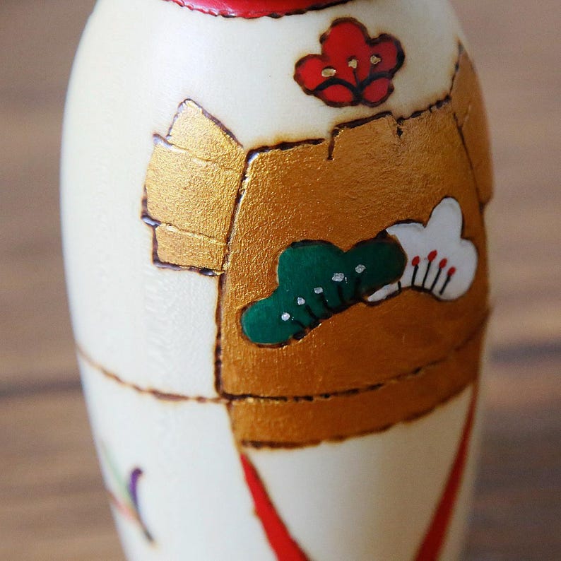 May include: A close-up of a Japanese Kokeshi doll. The doll is cream-colored with gold, red, and green painted details. The design includes a red flower, a green tree, and a white cloud with red accents. The doll has a red band around the top.