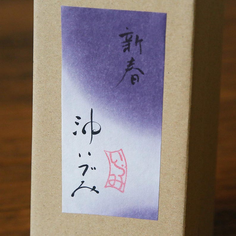 May include: A close-up of a brown cardboard box with a purple and white label. The label features Japanese calligraphy in black ink and a red stamp. The top text reads "New Spring".