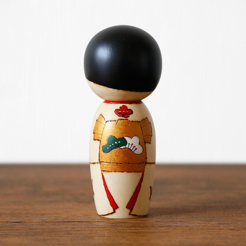 May include: A Japanese Kokeshi doll with a black head and a cream-colored body. The doll's body is decorated with gold, red, and green accents. The doll has a traditional design and is a collectible item.