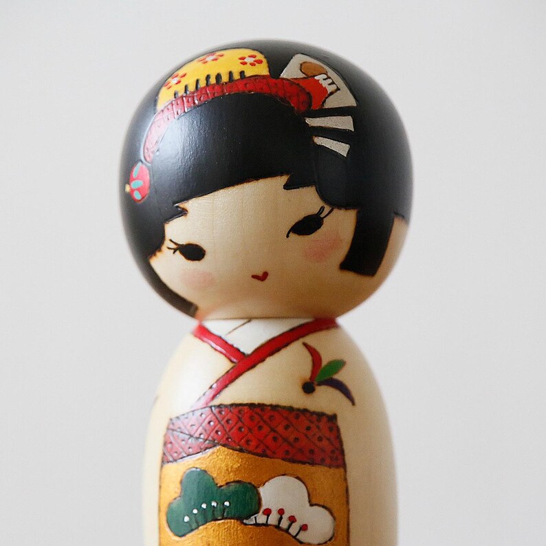 May include: A wooden Japanese Kokeshi doll with a black head and a painted face. The doll wears a cream-colored kimono with red and gold accents. The doll's hair is styled with a red and yellow floral design.