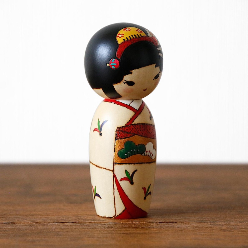 May include: A Japanese Kokeshi doll with a black head and a cream-colored body. The doll's head has a red and yellow headband. The body is decorated with red, green, and gold accents, and a red kimono-style design. The doll is standing on a wooden surface.