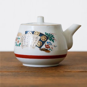May include: White ceramic teapot with a red band and a spout. The teapot is decorated with a fan design, flowers, and Japanese text. The lid has a small handle. The teapot is on a wooden surface.