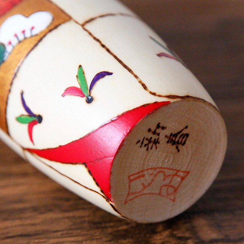 May include: A close-up of a Japanese Kokeshi doll. The doll is cylindrical with a cream-colored body, decorated with red, gold, green, and purple accents. The bottom of the doll has Japanese characters and a red stamp.
