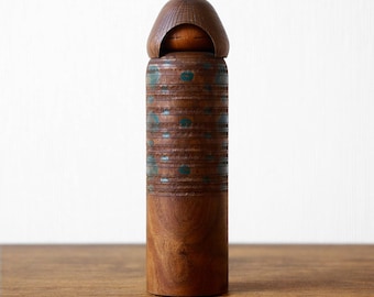 May include: A cylindrical wooden kokeshi doll featuring a smooth, rounded headpiece and a body decorated with horizontal carved bands and small, circular teal-coloured accents. The wood grain is visible throughout the natural brown surface.