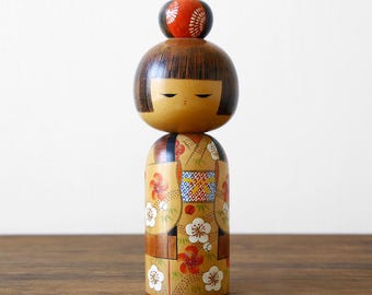 May include: A wooden Kokeshi doll featuring a rounded head with dark brown hair and closed eyes, topped with a small red circular ornament. The cylindrical body is painted in a light tan colour with a floral pattern of white and red blossoms.