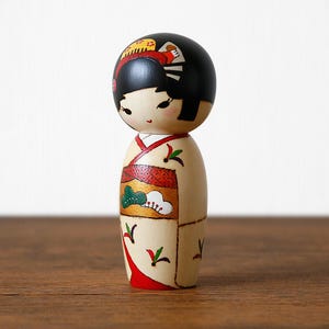 May include: A Japanese Kokeshi doll with a black head and a cream-colored body. The doll is wearing a kimono with red, green, and gold accents. The doll has black hair and a painted face with delicate features. The doll is standing on a wooden surface.