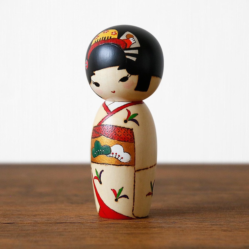 May include: A Japanese Kokeshi doll with a black head and a cream-colored body. The doll is wearing a kimono with red, green, and gold accents. The doll has black hair and a painted face with delicate features. The doll is standing on a wooden surface.