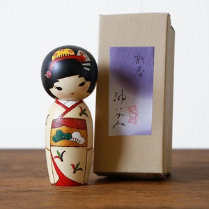 May include: A wooden Japanese Kokeshi doll with a black head and a cream-colored body. The doll is wearing a kimono with red and green accents. A cardboard box with Japanese text is next to the doll.