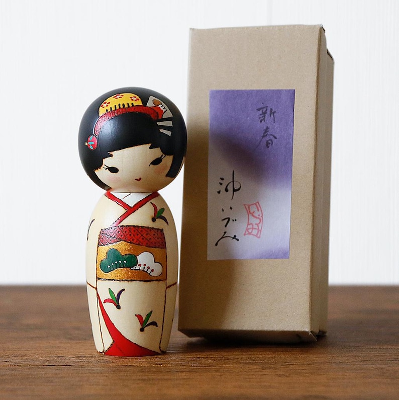 May include: A wooden Japanese Kokeshi doll with a black head and a cream-colored body. The doll is wearing a kimono with red and green accents. A cardboard box with Japanese text is next to the doll.