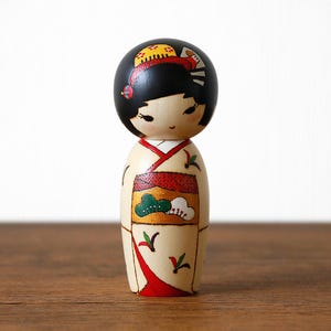 May include: A wooden Japanese Kokeshi doll with a black head and a cream-colored body. The doll is wearing a kimono with red, green, and gold accents. The doll has black hair and delicate facial features. The doll is standing on a wooden surface.