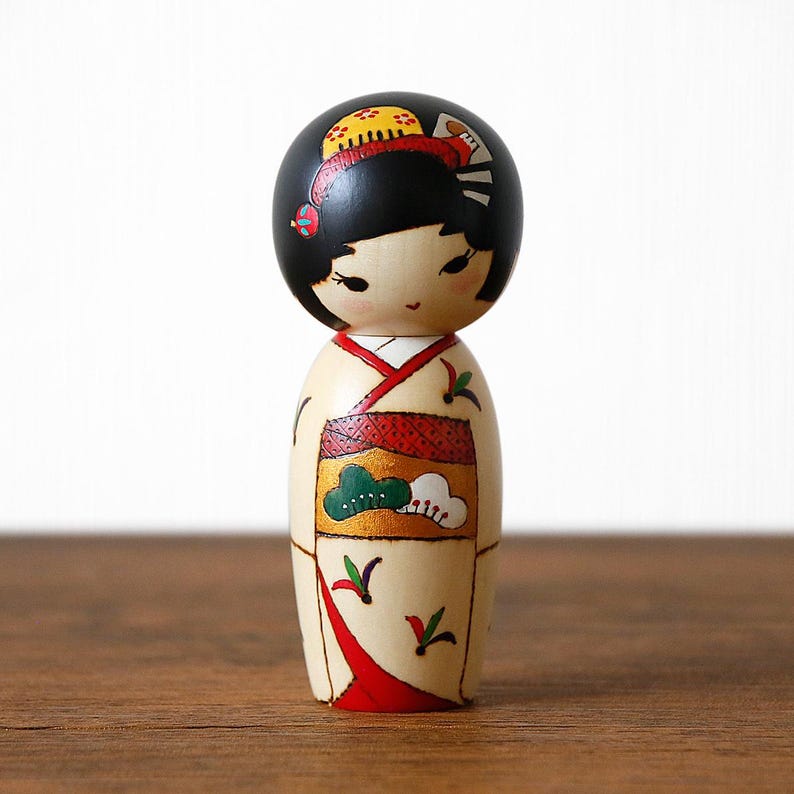 May include: A wooden Japanese Kokeshi doll with a black head and a cream-colored body. The doll is wearing a kimono with red, green, and gold accents. The doll has black hair and delicate facial features. The doll is standing on a wooden surface.
