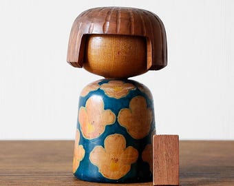May include: A wooden Kokeshi doll with a brown, carved hairstyle and a blue body decorated with orange floral patterns. A small, rectangular wooden block is beside the doll. The doll is a traditional Japanese craft.