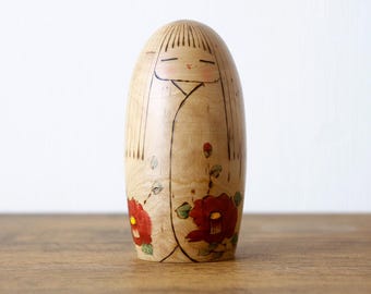 May include: A smooth, light-coloured wooden kokeshi doll with a rounded top. The doll features a hand-painted face with closed eyes and straight hair, and a kimono design decorated with red camellia flowers and green leaves on the lower body.