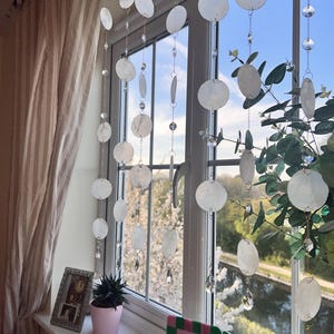 Könnte beinhalten: Dekorative Fensterdekoration mit Schnüren aus weißen, kreisförmigen Muschelschmuck und Kristallakzenten. Die Ornamente hängen vor einem Fenster mit Blick auf Bäume und blauen Himmel. Eine rosa Topfpflanze und ein gerahmtes Foto stehen auf der Fensterbank.